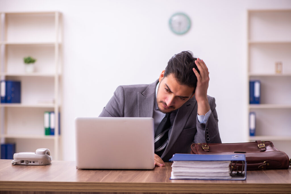 Man sitting at desk with an annoyed look in front of laptop with a compliance manual next to him.